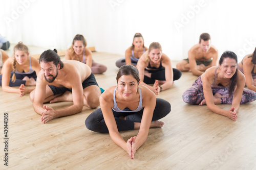 Group of young sporty attractive people in yoga studio, practicing yoga lesson with instructor, sitting on floor in forward band stretching yoga pose. Healthy active lifestyle, working out in gym