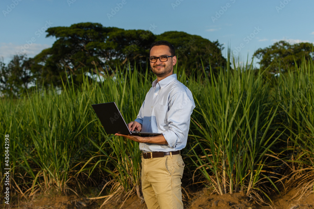 Formally dressed Latino man staring straight at the camera while holding his laptop posing in front of a sugarcane crop