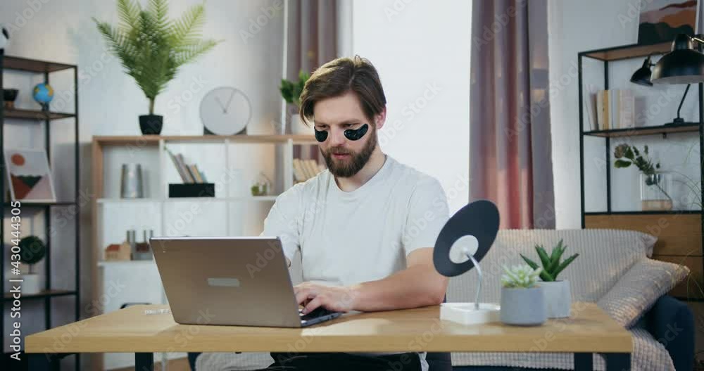 Good-looking smiling modern 30-aged bearder with anti-aging hydrogel eye-patches sitting at the table and working on computer,close up Good-looking smiling modern 30-aged bearder with anti-aging hydrogel eye-patches sitting at the table and working on computer,close up