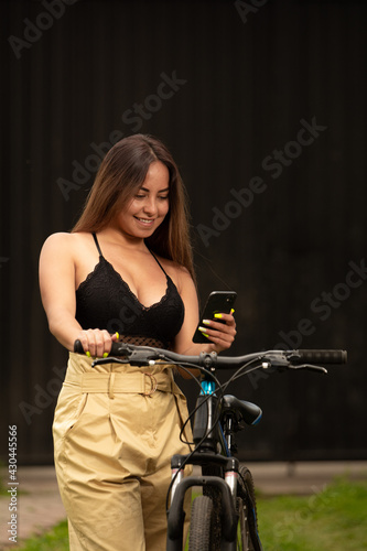 Smiling latin woman using a smart phone walking with a bicycle in an urban park