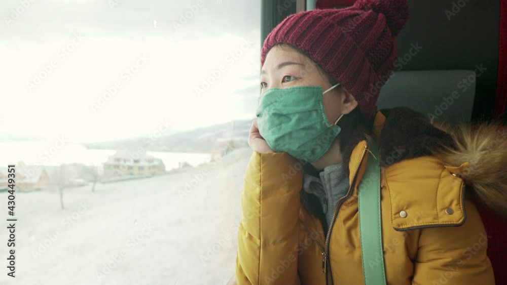train traveling in times of covid19 - young happy and beautiful Asian Korean woman in face mask looking snow landscape through railcar window during winter getaway