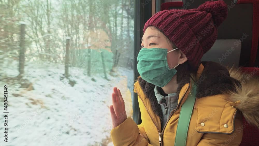 train traveling in times of covid19 - young happy and beautiful Asian Korean woman in face mask looking snow landscape through railcar window during winter getaway