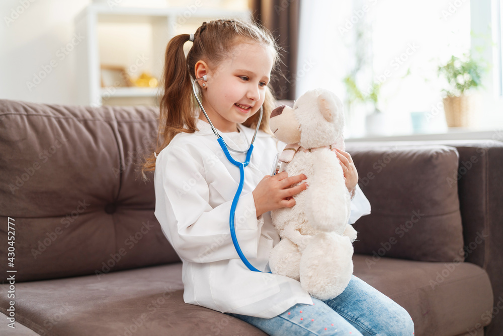 little Caucasian girl in glasses and a white medical uniform is treating a teddy bear, playing doctor and hospital. A happy child acts as a doctor examining a toy in a veterinary clinic. little Caucasian girl in glasses and a white medical uniform is treating a teddy bear, playing doctor and hospital. A happy child acts as a doctor examining a toy in a veterinary clinic.