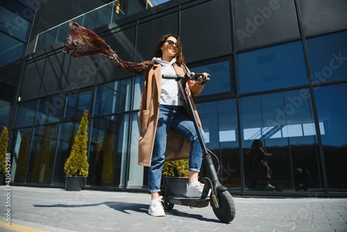 Young beautiful woman in a jacket smiles and rides an electric scooter to work along office buildings