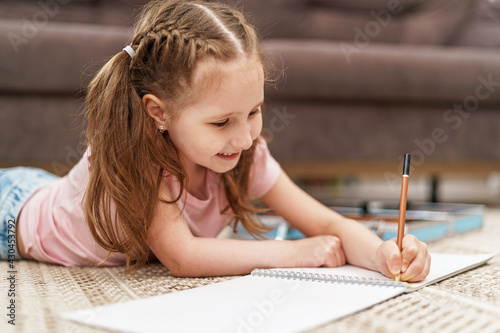 Little cute Caucasian girl child, drawing in an album with colored pencils, lying on floor. hardworking child does homework. The child's hobby is drawing. new modern set for drawing and creativity