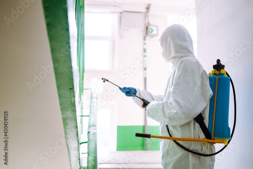 Man wearing protective suit disinfecting the entrance of a residential building with spray chemicals to preventing the spread of coronavirus, pandemic in quarantine city. Covid -19. Cleaning concept. © maxbelchenko