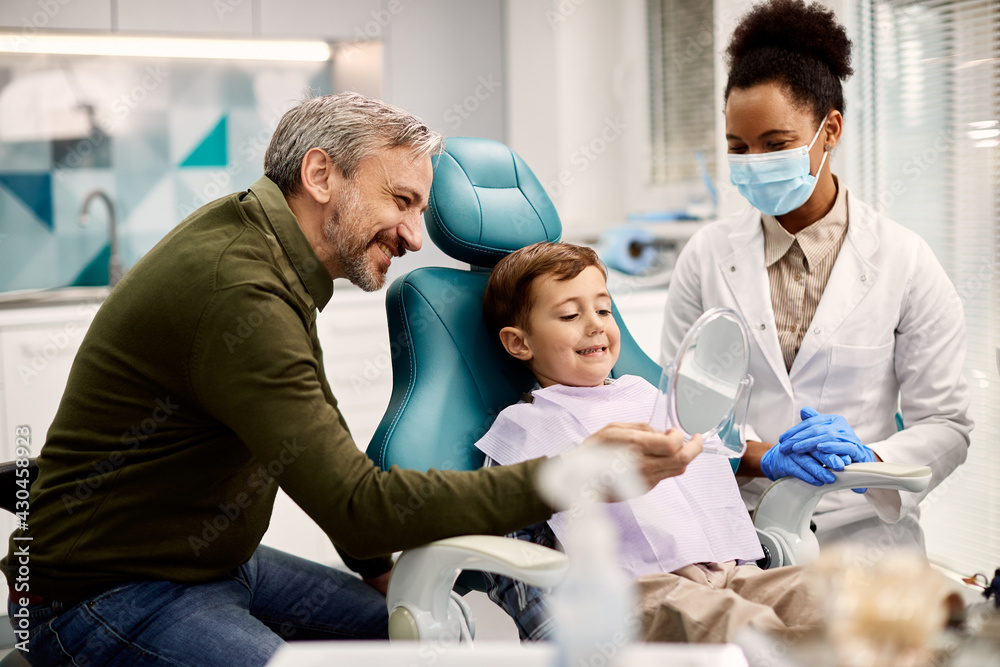 Obraz premium Happy boy looking at his teeth in a mirror while being with his father at dentist's office.
