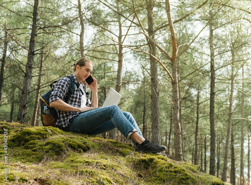 Young man sitting on ground at forest talking on smartphone and working on laptop