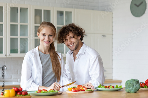 Happy couple standing in kitchen at home preparing together fresh vegetable salad.