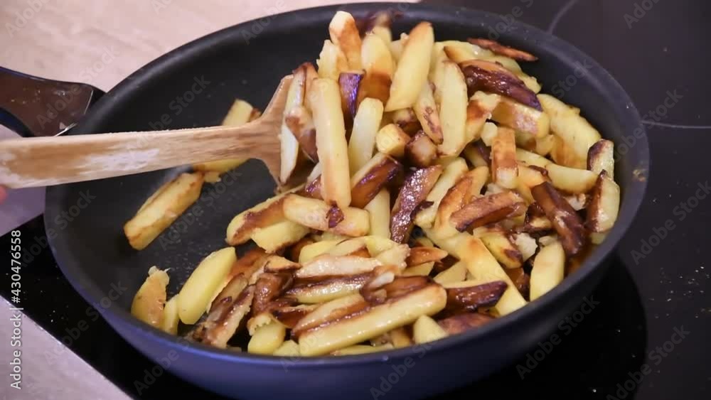 Hand with wooden paddle stir homemade French fries in frying pan at kitchen stove. Cooking of crispy fried potatoes with curry. Using frying spatula to mix hot dish. Slow motion