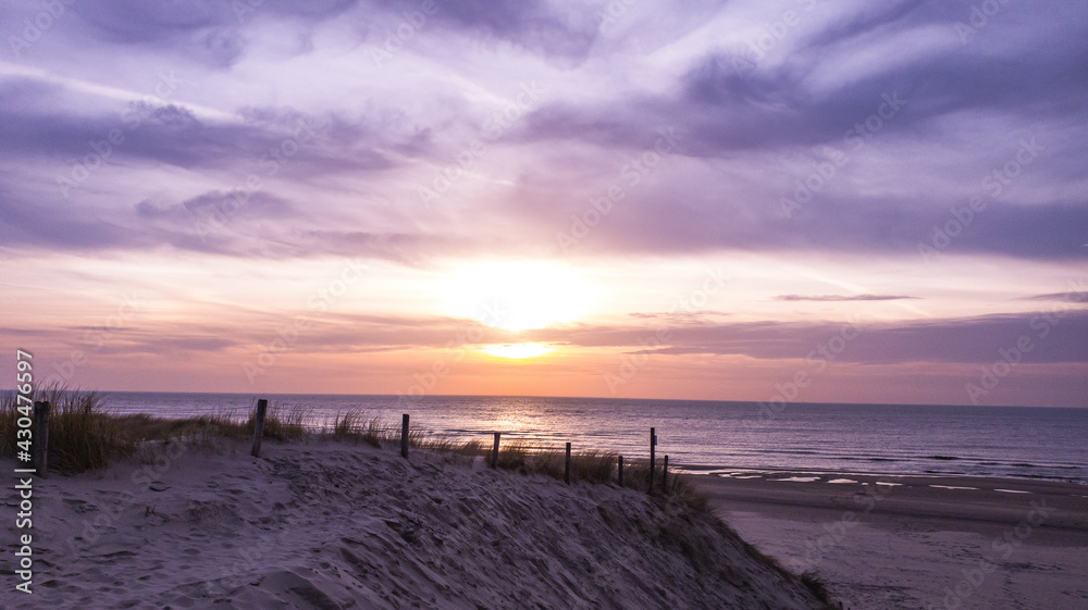 Obraz premium Dramatic sunset over viewing the beach from the dunes, the sunlight is reflected by the sea water and the sun is surrounded by dramatic clouds