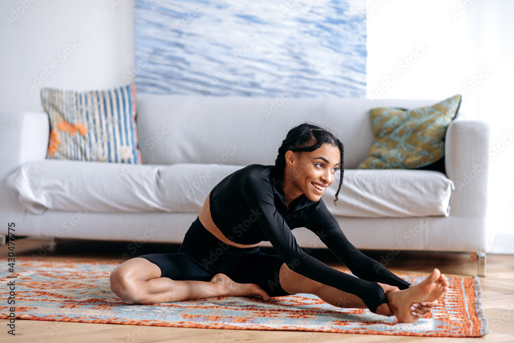 Fototapeta premium Satisfied young African American woman in black sportswear, doing fitness in living room on carpet, stretching her arms to her feet, doing back stretching, leads healthy lifestyle, looking away, smile