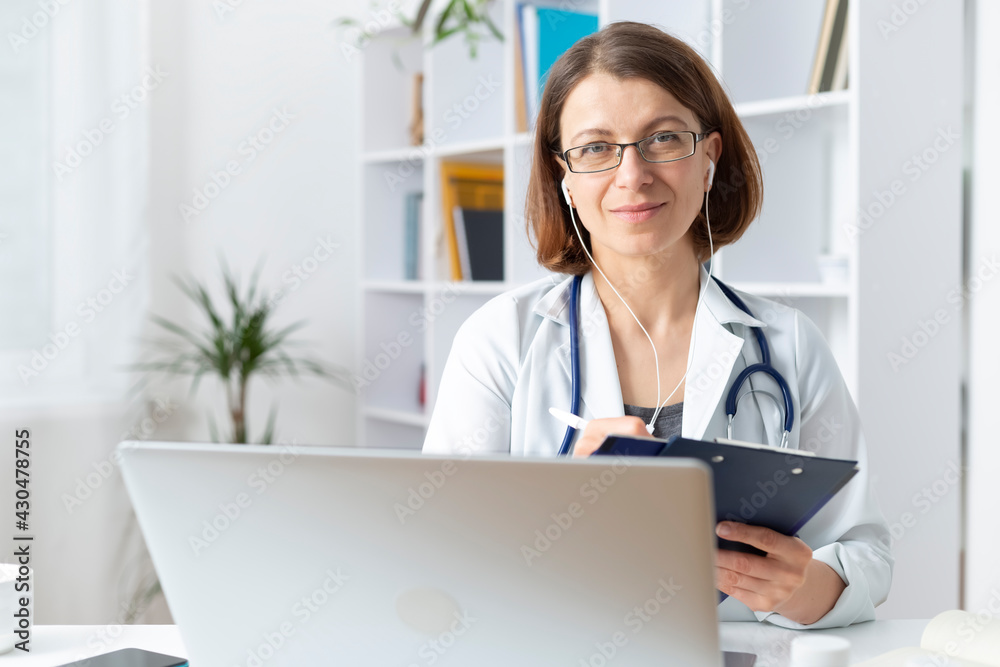 Portrait of a woman doctor in the interior working with a laptop, wearing headphones, consulting remotely.