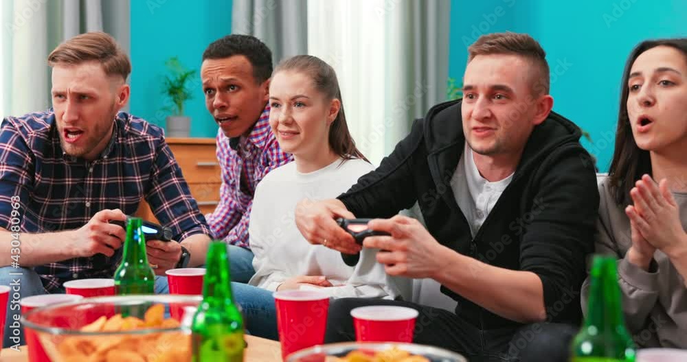 Cheerful mixed races friends resting in room sitting on sfoa playing video games using joystick. Friends having together at home. Gamers concept. Home party Cheerful mixed races friends resting in room sitting on sfoa playing video games using joystick. Friends having together at home. Gamers concept. Home party