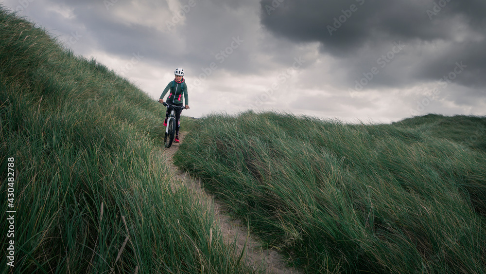 Woman with bike in sand dunes with grass at coast in Denmark, dark clouds in dramatic sky.