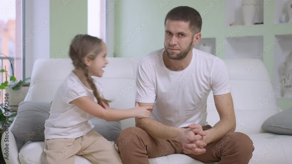 Playful Caucasian girl pulling hand of tired young father in slow motion sitting on couch at home. Positive Caucasian man looking at camera with exhausted facial expression and grimacing with daughter Playful Caucasian girl pulling hand of tired young father in slow motion sitting on couch at home. Positive Caucasian man looking at camera with exhausted facial expression and grimacing with daughter
