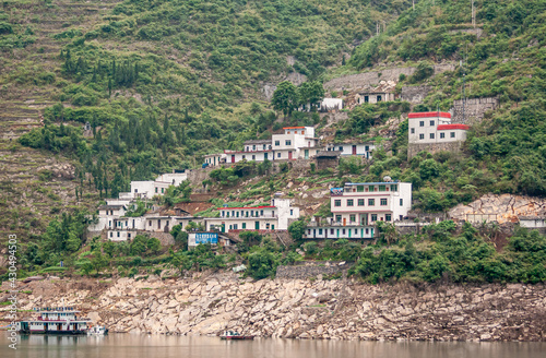 Guandukou, Hubei, China - May 7, 2010: Wu Gorge on Yangtze River. White buildings with colroed trims sit above each other on slope of green forested mountain. Boats on brownish water.