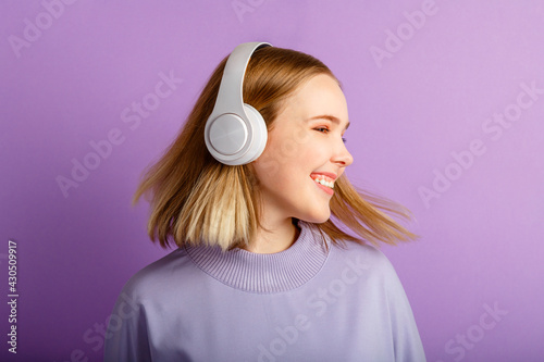 Attractive smiling woman dancing in headphones with flying blonde hair hairstyle. Teenager girl portrait looking side enjoy listen music moving in headphones isolated over purple color background.