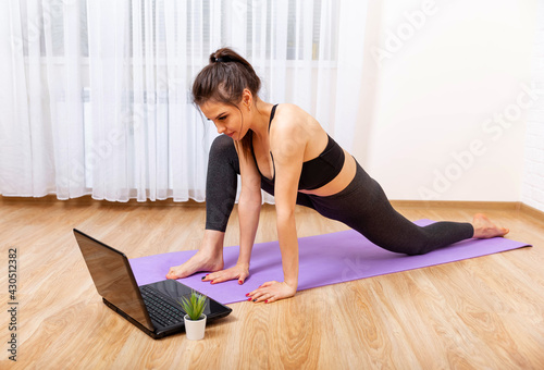 Fit woman doing yoga and watching online tutorials on laptop. Attractive young woman doing yoga and stretching online at home.Girl doing yoga at home. Yoga online on the laptop. Healthy lifestyle