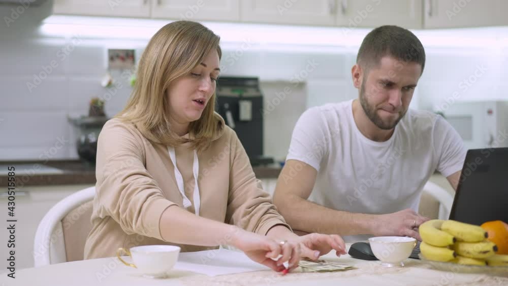 Caucasian woman counting money talking with blurred man surfing Internet on laptop at background. Wife dissatisfied with husband income sitting in kitchen at home. Freelance and relationship Caucasian woman counting money talking with blurred man surfing Internet on laptop at background. Wife dissatisfied with husband income sitting in kitchen at home. Freelance and relationship