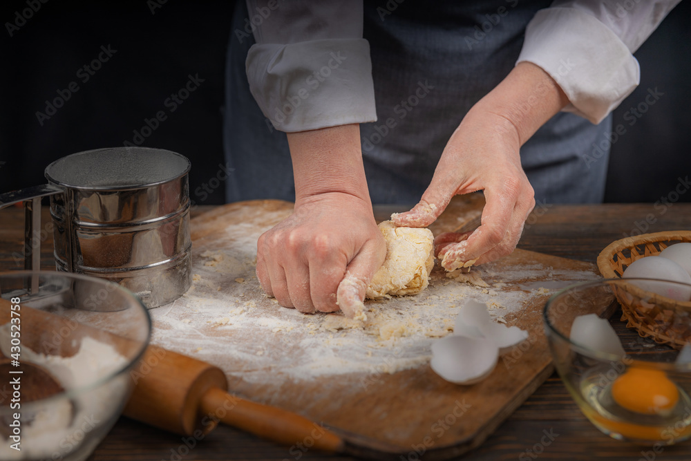 Women's hands, flour and dough. A woman in an apron cooking dough for homemade baking, a rustic home cozy atmosphere, a dark background with unusual lighting. Women's hands, flour and dough. A woman in an apron cooking dough for homemade baking, a rustic home cozy atmosphere, a dark background with unusual lighting.