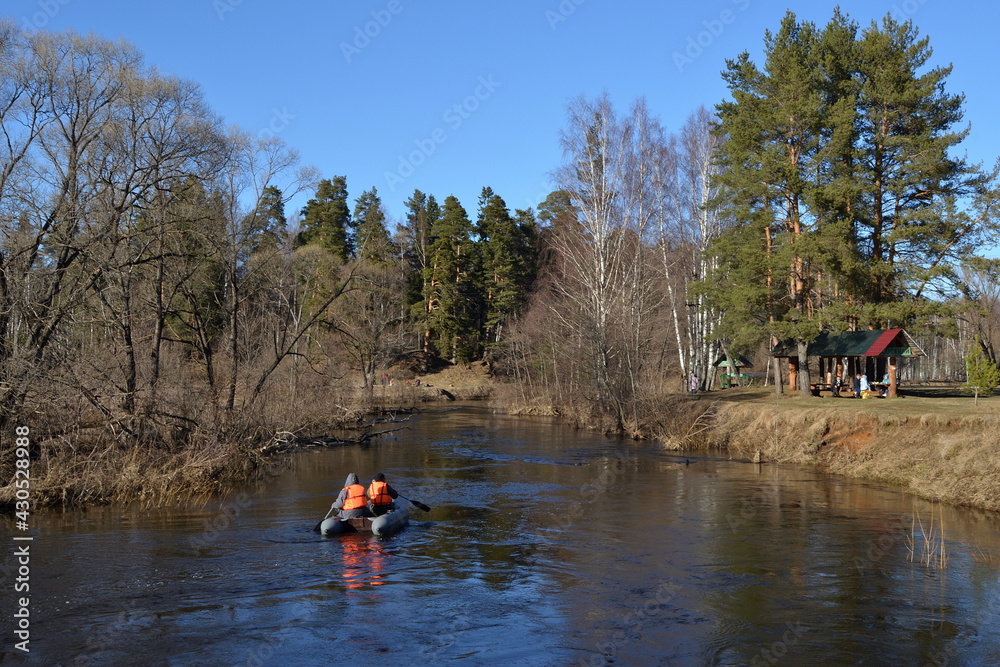 Kayaking on the river in the spring. A kayak with two athletes floats down the river on a sunny spring day, green trees, bright blue sky. People have picnics on the banks of the river.