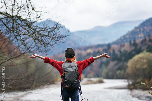 a traveler in a hat  sweater and trousers gestures with her hands on the river bank in the mountains