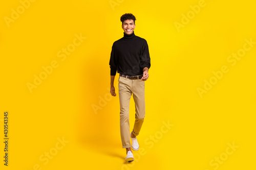 Full size photo of optimistic brunet guy go wear black sweater trousers sneakers isolated on yellow color background