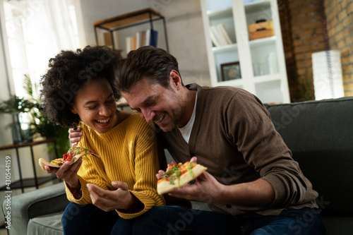  Cheerful young couple sitting on sofa at home. Happy woman and man eating pizza while watching a movie