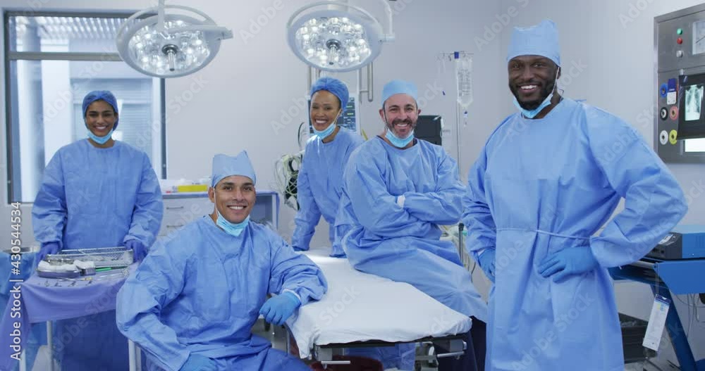 Diverse male and female doctors wearing face masks standing in operating theatre smiling to camera Diverse male and female doctors wearing face masks standing in operating theatre smiling to camera