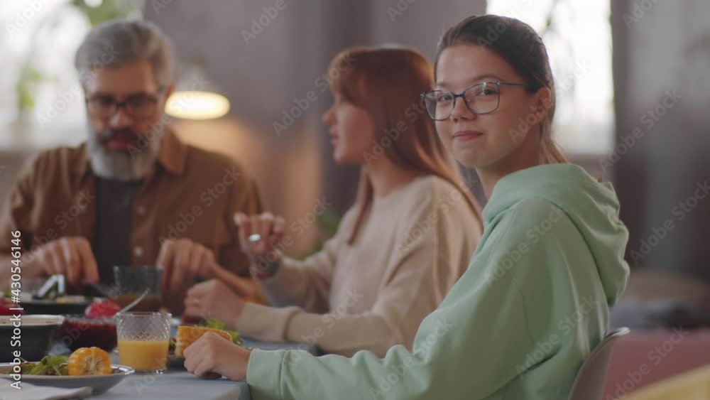 Portrait of pretty teen girl in glasses and hoodie sitting at dinner table with family, looking at camera and smiling