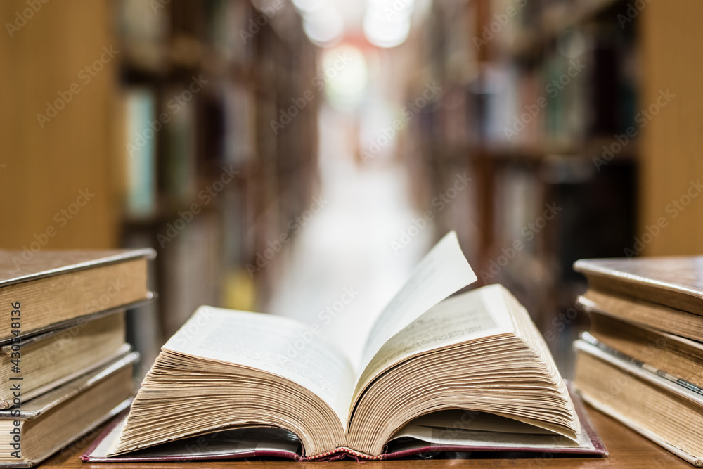 Old book in library with open textbook, stack piles of literature text archive on reading desk, and blur aisle of bookshelves in school study class room background for education learning concept Old book in library with open textbook, stack piles of literature text archive on reading desk, and blur aisle of bookshelves in school study class room background for education learning concept