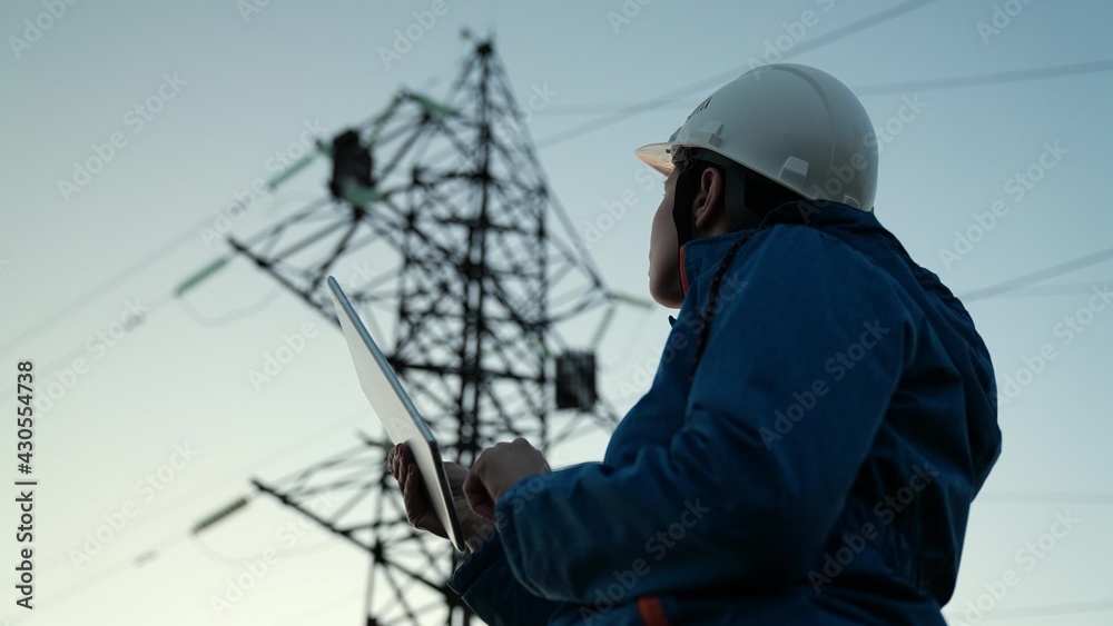 Woman power engineer in white helmet checks power line using computer on tablet, remote control of power system. High voltage electrical lines at sunset. Distribution and supply of electricity. Energy