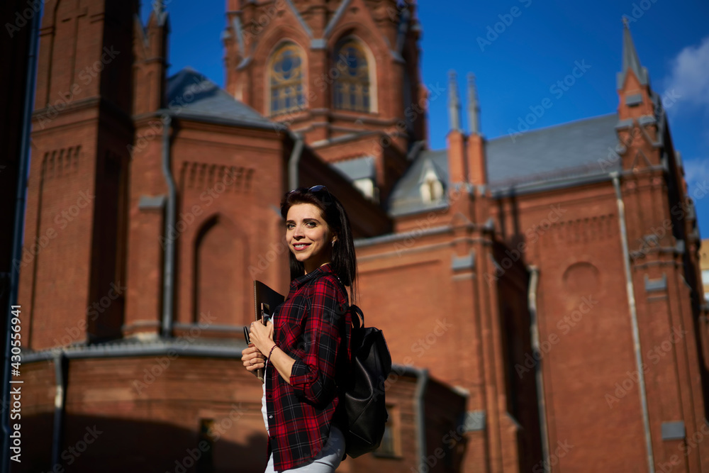 Half length portrait of cheerful Caucasian woman in casual clothes smiling at camera while exploring touristic town during summer vacations,happy female enjoying sunny weather during walk time in city