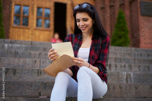 Pretty millennial student with yellow envelope reading received mail notification during daytime in city, Caucasian hipster girl checking message and correspondence holding postal paper and postcard