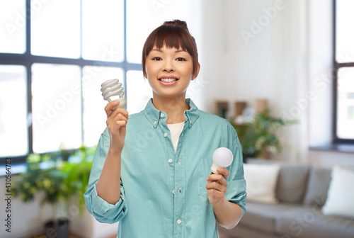 eco living  inspiration and sustainability concept - portrait of happy smiling young asian woman in turquoise shirt holding energy saving lighting bulb over home room background