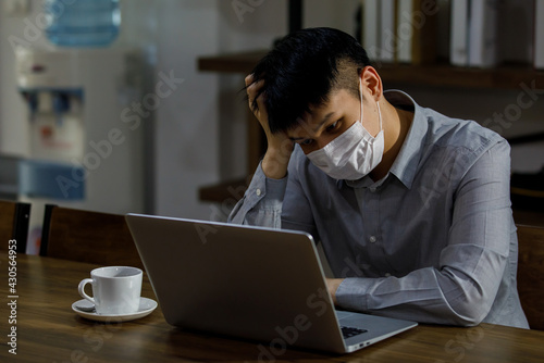 young Asian businessman wearing gray shirt and face mask sitting in the office looking at a laptop computer screen thinking and seriously while working and communicate online