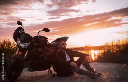 Young handsome male biker, is sitting on the ground near his motorcycle and watching on the sunset.