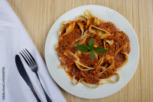 Pasta with bolognese tomato sauce and basil leaves served on a white plate in the background, bamboo mat and napkin with fork and knife beside it. top view photo.
