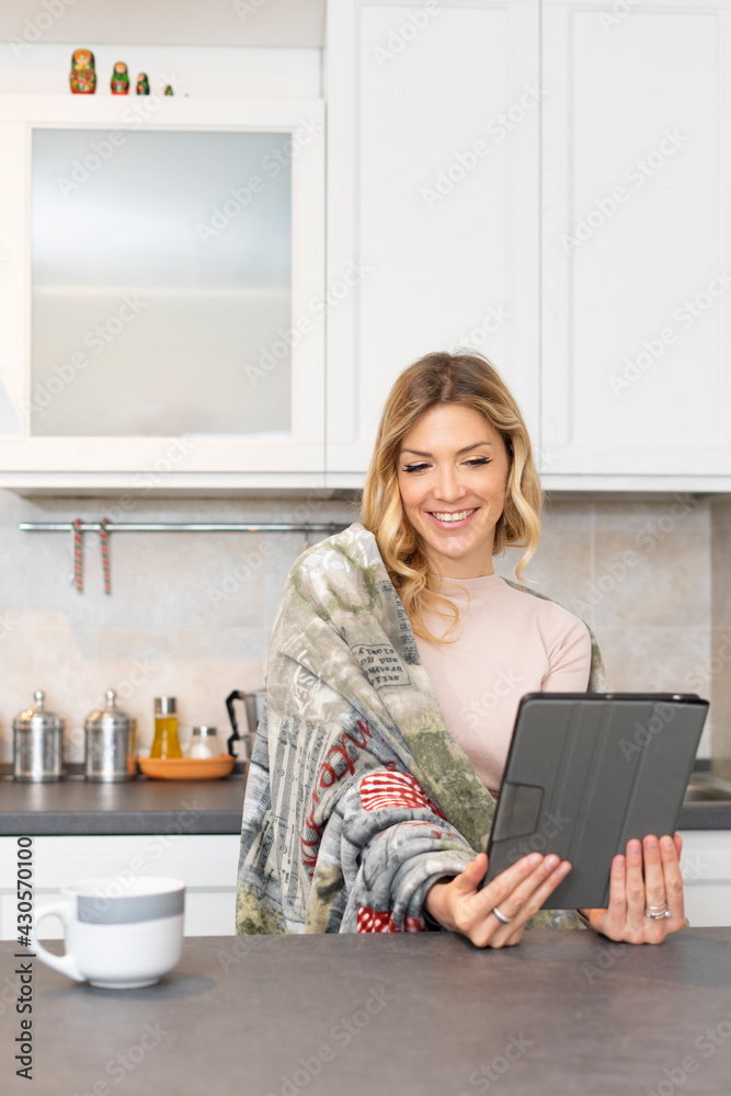 Beautiful smiling woman enjoys a video call. Blonde girl at home holding a tablet in the kitchen. Woman sitting with shoulders covered with a warm blanket. Beautiful smiling woman enjoys a video call. Blonde girl at home holding a tablet in the kitchen. Woman sitting with shoulders covered with a warm blanket.
