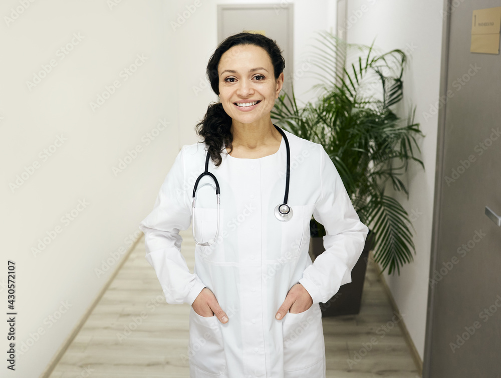 General practitioner. Smiling female mixed-race physician wearing a medical uniform with a stethoscope stands in a hospital corridor General practitioner. Smiling female mixed-race physician wearing a medical uniform with a stethoscope stands in a hospital corridor