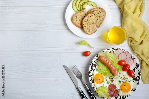 Plate with scrambled eggs, sausages, fresh tomatoes and salad, avocado on a light wooden background, top view. Layout, copy space. Delicious and nutritious breakfast