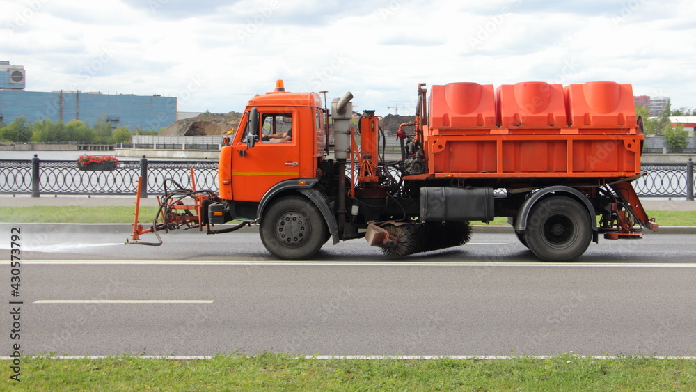 New Russian orange street sweeper truck pours asphalt road, street washing in Moscow, city improvement by municipal services on a sunny summer day
