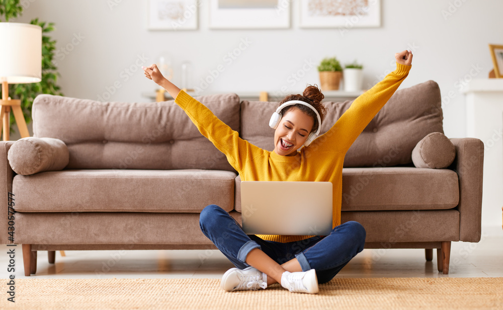 Overjoyed african american female in headphones sitting on floor and celebrating success while working remotely or studying online - obrazy, fototapety, plakaty 