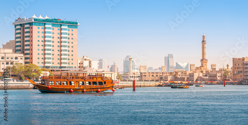 Photography An authentic wooden cruise ship cruises and transports tourists along the Dubai Creek Canal with skyscrapers and minarets of mosques in the background