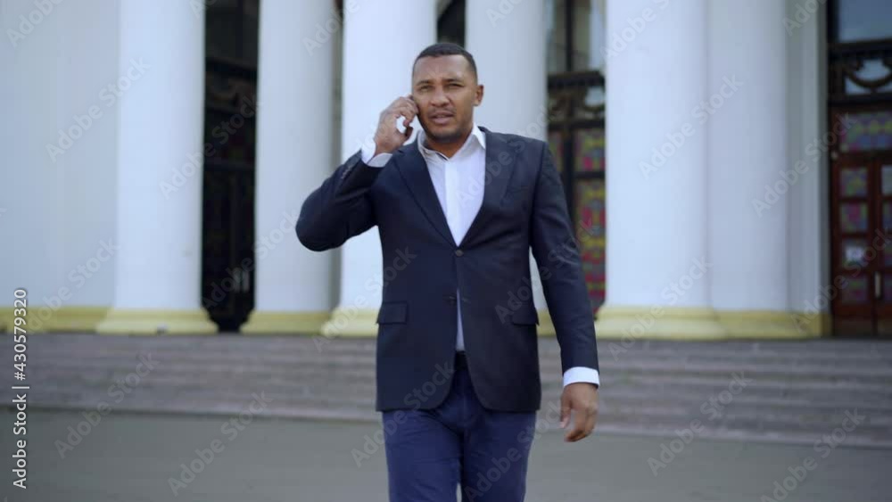 Middle shot of concentrated successful businessman talking on the phone walking outdoors with building with columns at background. Portrait of confident African American man in suit
