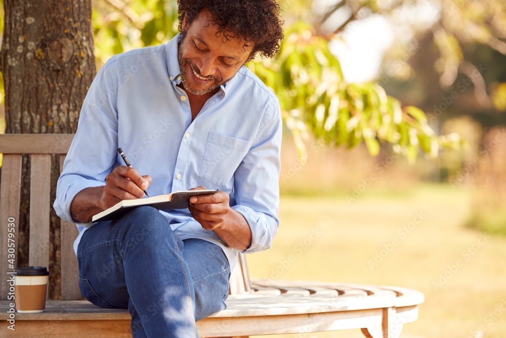 Fototapeta premium Mature Man With Takeaway Coffee Sitting On Park Bench Under Tree Writing In Notebook