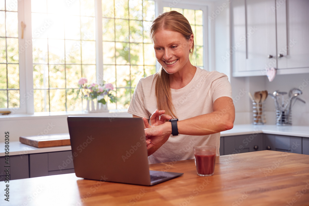 Mature Woman Wearing Fitness Clothing At Home Logging Activity From Smart Watch Onto Laptop Mature Woman Wearing Fitness Clothing At Home Logging Activity From Smart Watch Onto Laptop