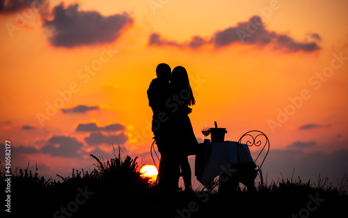 Couple is celebrating love at sunset. Close couple is hugging and kissing with table and chairs and bottle of champagne outdoors at sunset. 