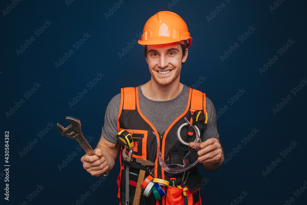 Smiling builder in a helmet is holding glasses and wrench, isolated over blue background Smiling builder in a helmet is holding glasses and wrench, isolated over blue background